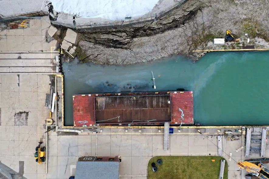 Aerial view of collapsed dock that has spilled gravel into a boat slip. An oily sheen is visible on the water.